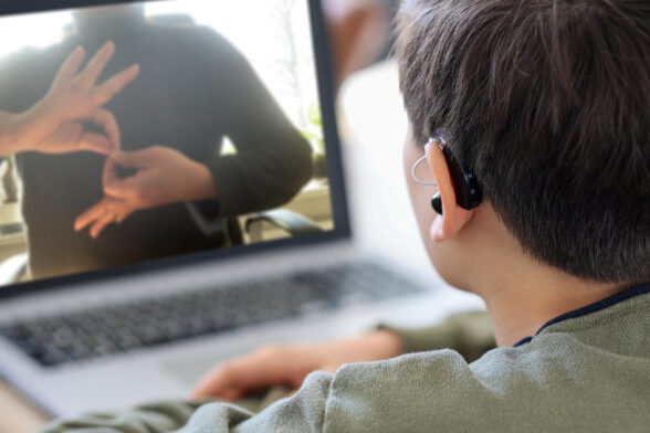 Photo d'un garçon qui porte un appareil auditif, qui regarde un écran d'ordinateur où une personne signe. - Agrandir l'image, fenêtre modale