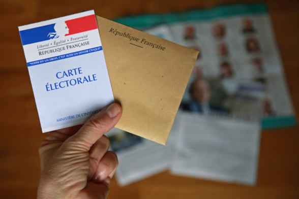 Photo d'une main qui tient une carte électorale et une enveloppe au dessus des flyers des candidats. - Agrandir l'image, fenêtre modale